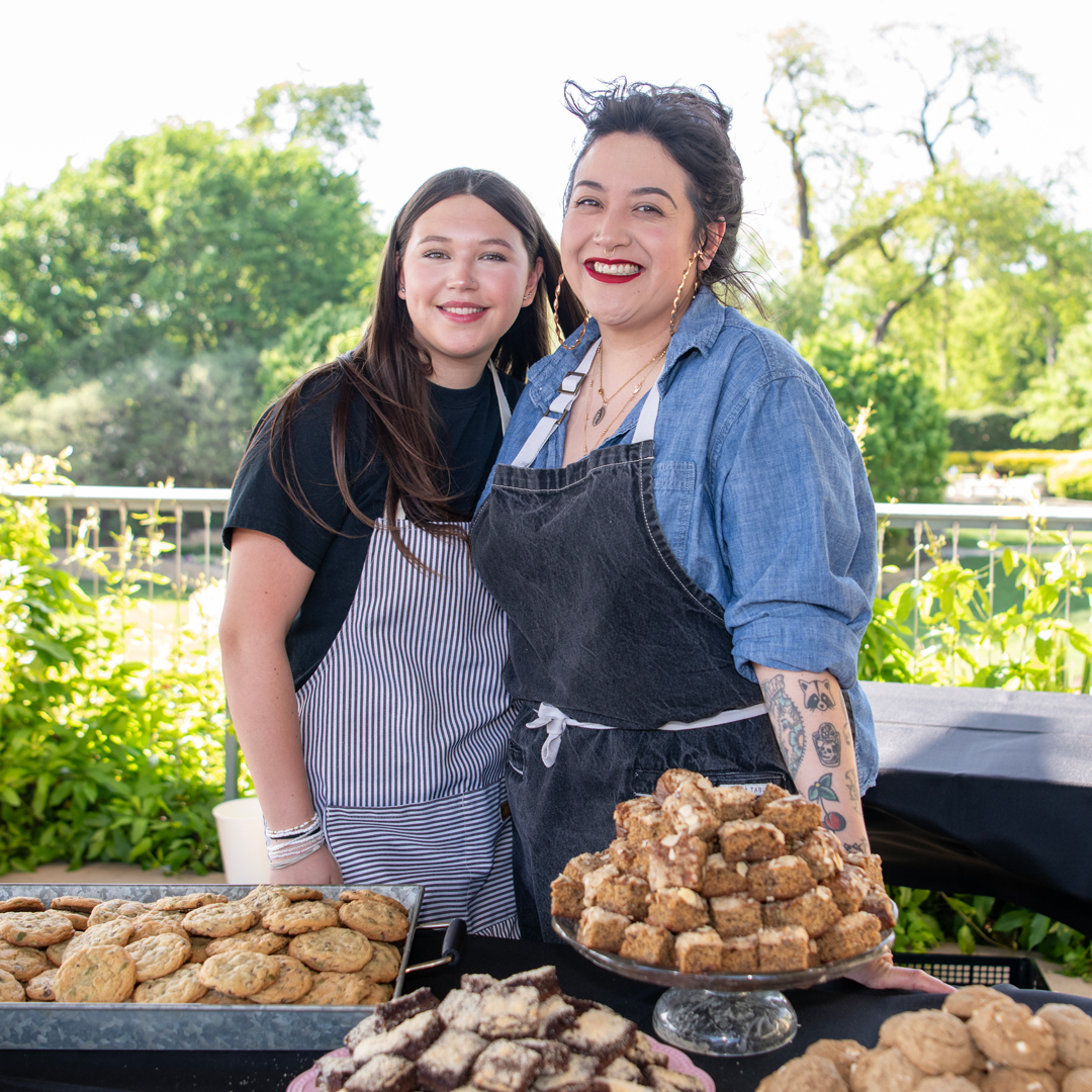 Two chefs standing behind their table at 2025 Bits and Bites event with a table of baked goods in front of them