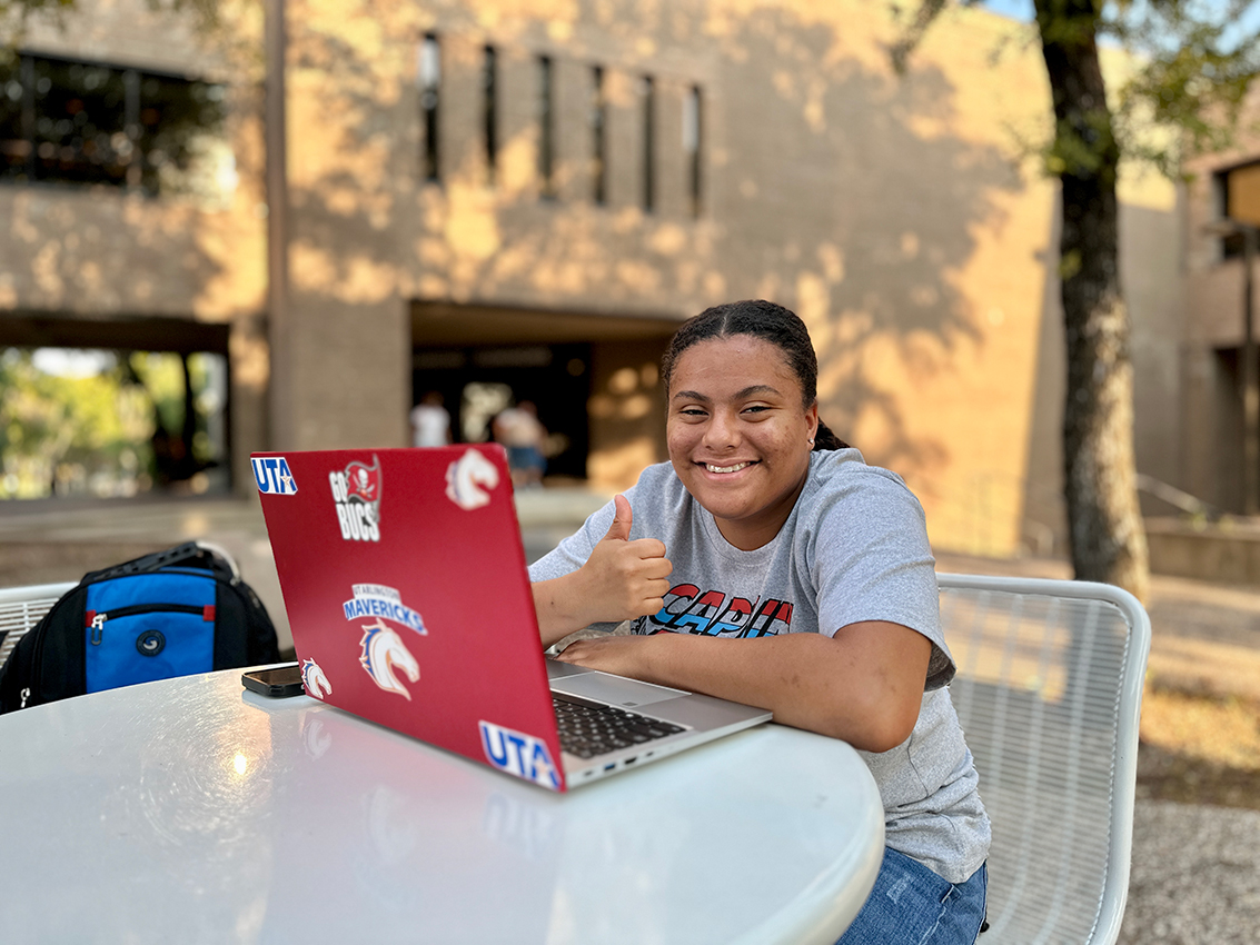 Student with laptop working outside on Dallas College campus smiling with thumbs up